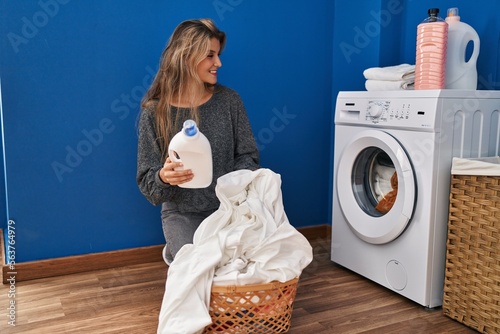 Young blonde woman smiling confident holding detergent bottle at laundry room