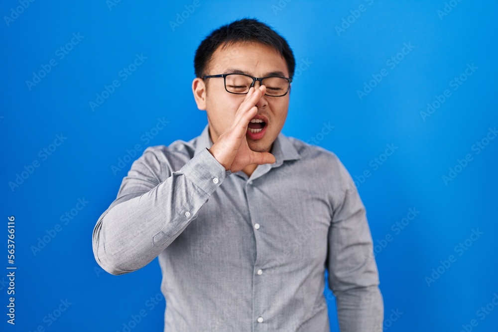 Young chinese man standing over blue background shouting and screaming loud to side with hand on mouth. communication concept.