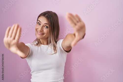 Blonde caucasian woman standing over pink background doing frame using hands palms and fingers  camera perspective