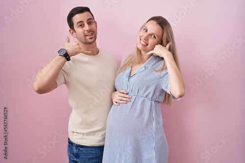 Young couple expecting a baby standing over pink background smiling doing phone gesture with hand and fingers like talking on the telephone. communicating concepts.