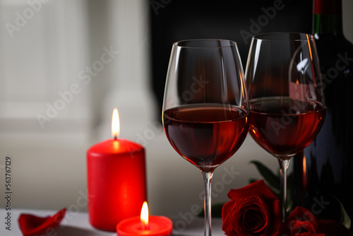 Glasses of red wine, burning candles and rose flowers against blurred background, space for text. Romantic atmosphere
