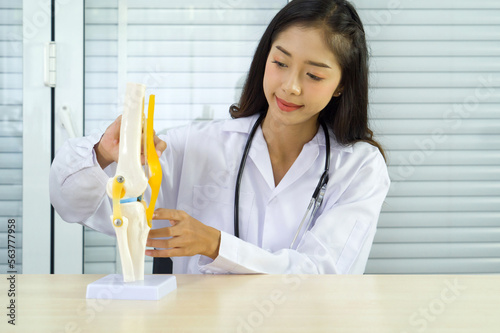 Schilderij op canvas Young asian doctor in white gown and stethoscope pointing at anterior cruciate ligament on deluxe functional knee joint model