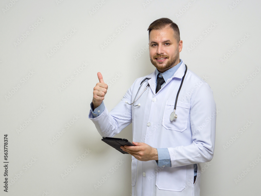Young caucasian doctor with short hair, mustache and beard dressed in white gown and stethoscope raise finger thumb up while holding tablet computer. Stand in front of white wall. Young caucasian doctor with short hair, mustache and beard dressed in white gown and stethoscope raise finger thumb up while holding tablet computer. Stand in front of white wall.