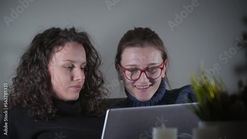 Two young beautiful female hiccup and smiling student with laptop sitting on sofa in an appartment photo