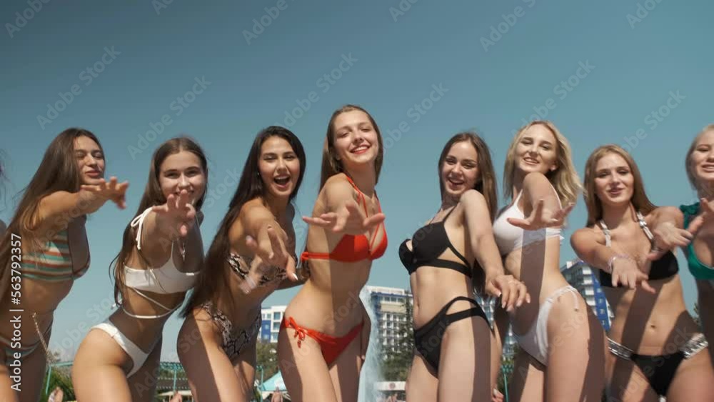 Happy young girls in swimsuit waving at camera near pool in water park