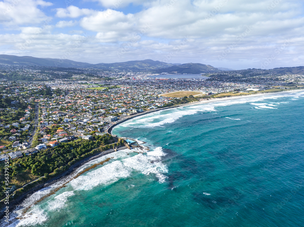 High angle aerial drone view of St Clair, a beachside suburb of Dunedin, second-largest city in the South Island of New Zealand. Dunedin city in the background. Hot water salt rock pool in foreground.
