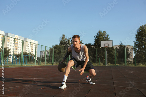 Athletic man playing basketball and dribbling ball on urban court in city during training in summer 