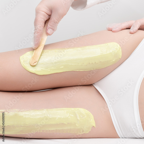 Cosmetologist in gloves applies green hot wax on slim woman legs using spatula while woman lying down on couch. Depilation with hot wax in beauty salon. Close up side view. Part of photo series.
