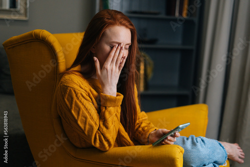 Portrait of tired young woman having blurry vision is rubbing and squinting eyes while typing using smartphone. Bored exhausted female checking phone in night sitting in yellow armchair at home.