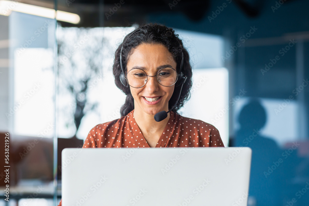 Smiling hispanic businesswoman working inside office with laptop and headset for video call, woman sitting at workplace happy working with clients. - obrazy, fototapety, plakaty 