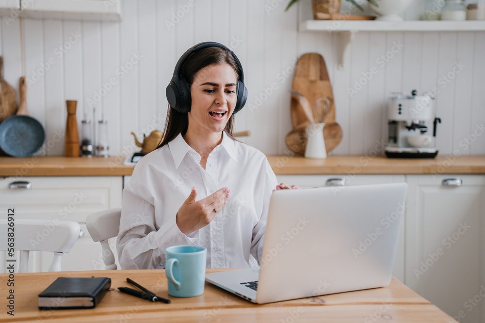 Purposeful brunette Hispanic girl in white shirt sitting at desk with laptop using headphones learns foreign language via internet at home. Cheerful young woman discussing with friend at video call. Purposeful brunette Hispanic girl in white shirt sitting at desk with laptop using headphones learns foreign language via internet at home. Cheerful young woman discussing with friend at video call.
