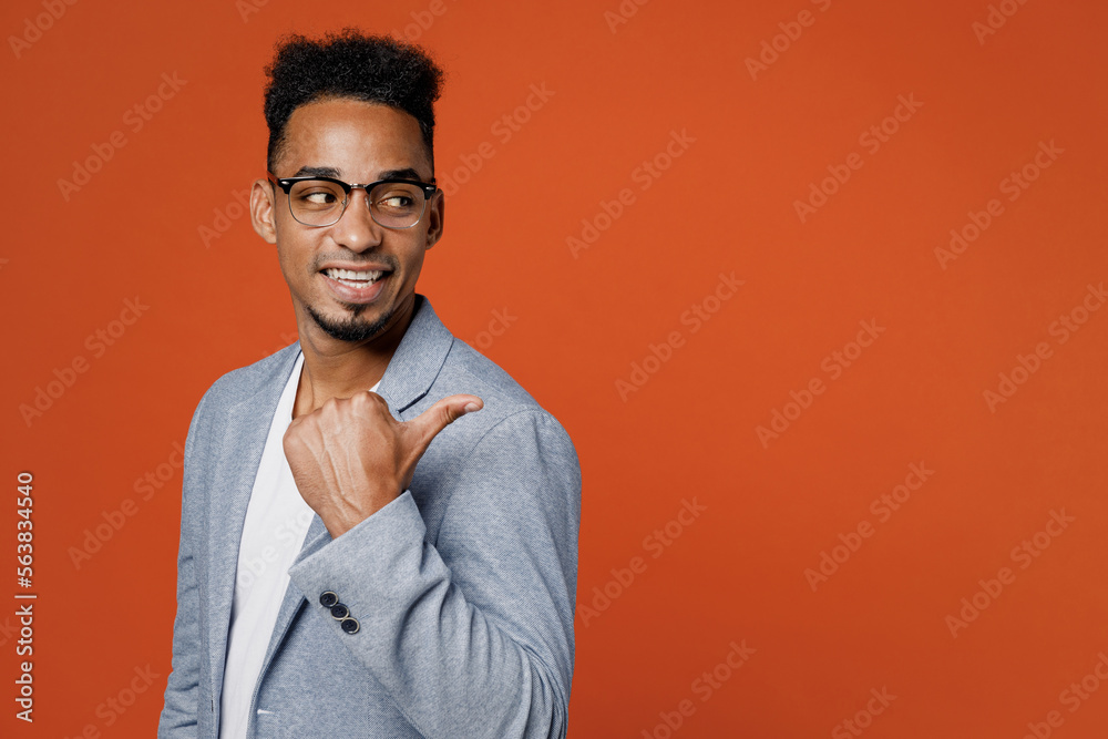 Side view fun young employee business man corporate lawyer wear classic formal grey suit shirt glasses work in office point thumb finger aside on area isolated on plain red orange background studio. Side view fun young employee business man corporate lawyer wear classic formal grey suit shirt glasses work in office point thumb finger aside on area isolated on plain red orange background studio.