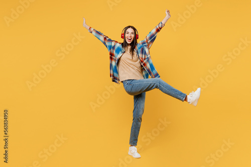 Full body young woman wear blue shirt beige t-shirt headphones listen to music dance with outstretched hands raise up leg isolated on plain yellow background studio portrait. People lifestyle concept.