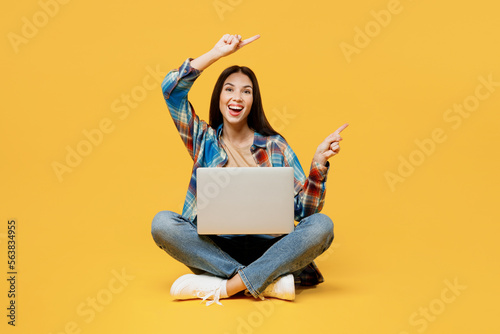 Full body young smiling cheerful happy smart IT woman wear blue shirt beige t-shirt sit hold use work on laptop pc computer point index finger aside on area isolated on plain yellow background studio.