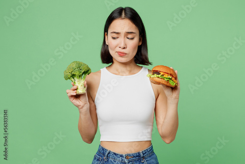 Young minded thoughtful sad woman wear white clothes hold burger broccoli count calories isolated on plain pastel light green background. Proper nutrition healthy fast food unhealthy choice concept.