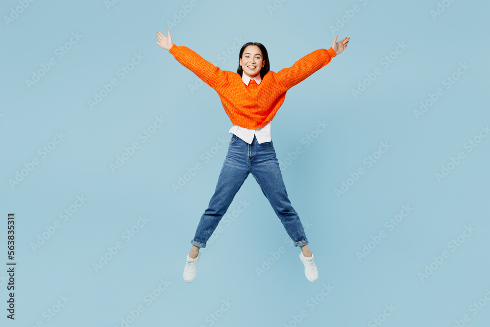Full body happy young woman of Asian ethnicity wear orange sweater glasses jump high with outstretched hands legs isolated on plain pastel light blue cyan background studio. People lifestyle concept. Full body happy young woman of Asian ethnicity wear orange sweater glasses jump high with outstretched hands legs isolated on plain pastel light blue cyan background studio. People lifestyle concept.