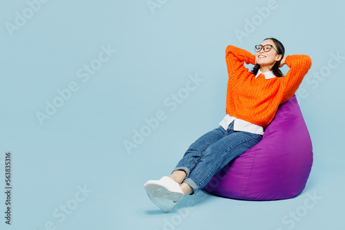 Full body smiling young woman of Asian ethnicity in orange sweater glasses sit in bag chair hold hands behind neck isolated on plain pastel light blue cyan background studio People lifestyle concept