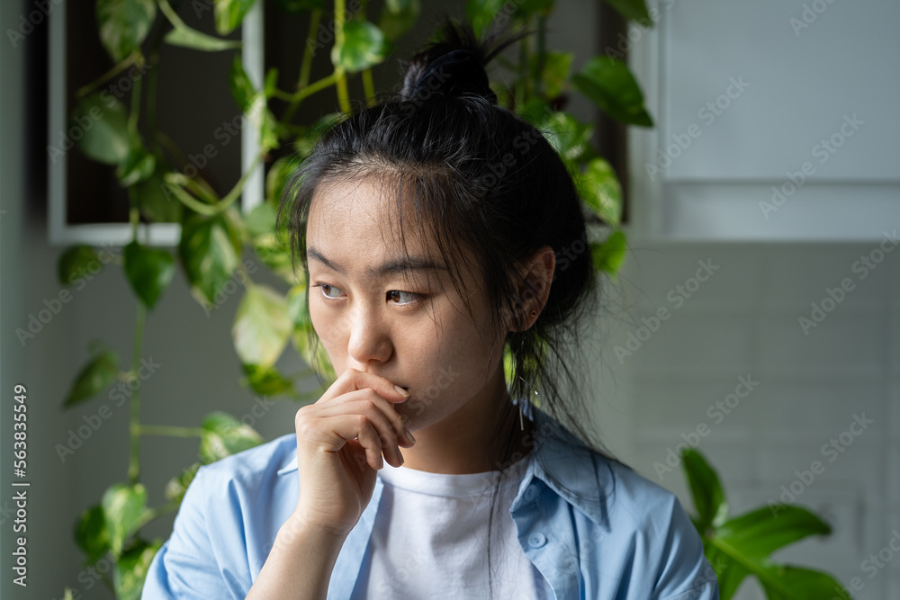 Thoughtful melancholic Asian woman touches lips with hand experiencing psychological disorder. Unhappy Chinese female stands in room near closet with houseplants and needs help of psychotherapist. Thoughtful melancholic Asian woman touches lips with hand experiencing psychological disorder. Unhappy Chinese female stands in room near closet with houseplants and needs help of psychotherapist.