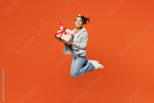 Full body side view young woman of African American ethnicity she wear grey shirt headband jump high hold present box with gift ribbon bow isolated on plain orange background People lifestyle concept