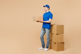 Full body sideways delivery guy employee man wear blue cap t-shirt uniform workwear work as dealer courier hold cardboard box stand near stack isolated on plain light beige background Service concept