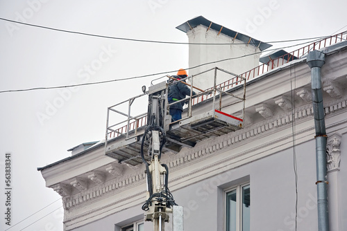 Man on lifting platform remove icicles and clean snow from building roof. Man breaking icicles on rooftop, clearing snow from roof of an old building. Worker on lift platform remove ice at height