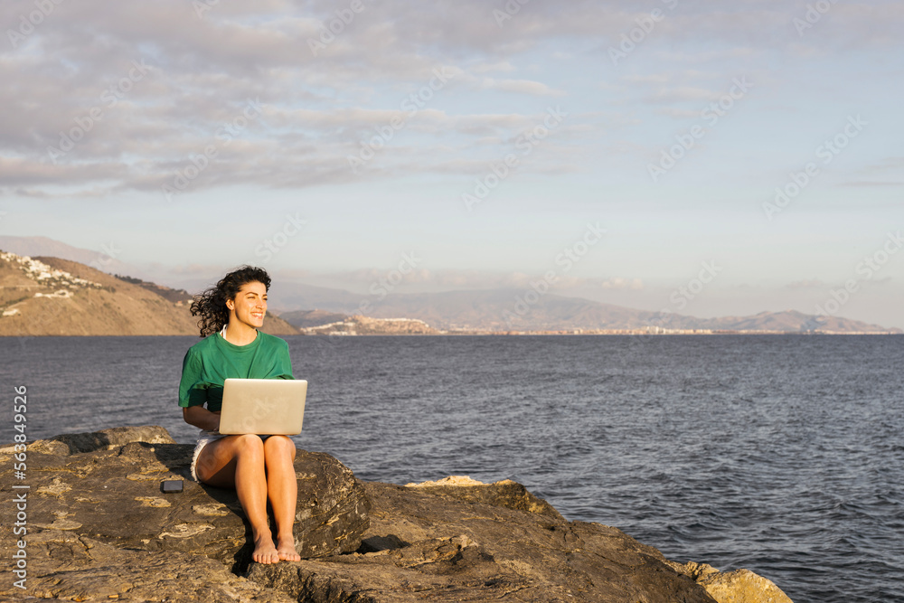Smiling woman with laptop sitting on rock by sea Smiling woman with laptop sitting on rock by sea