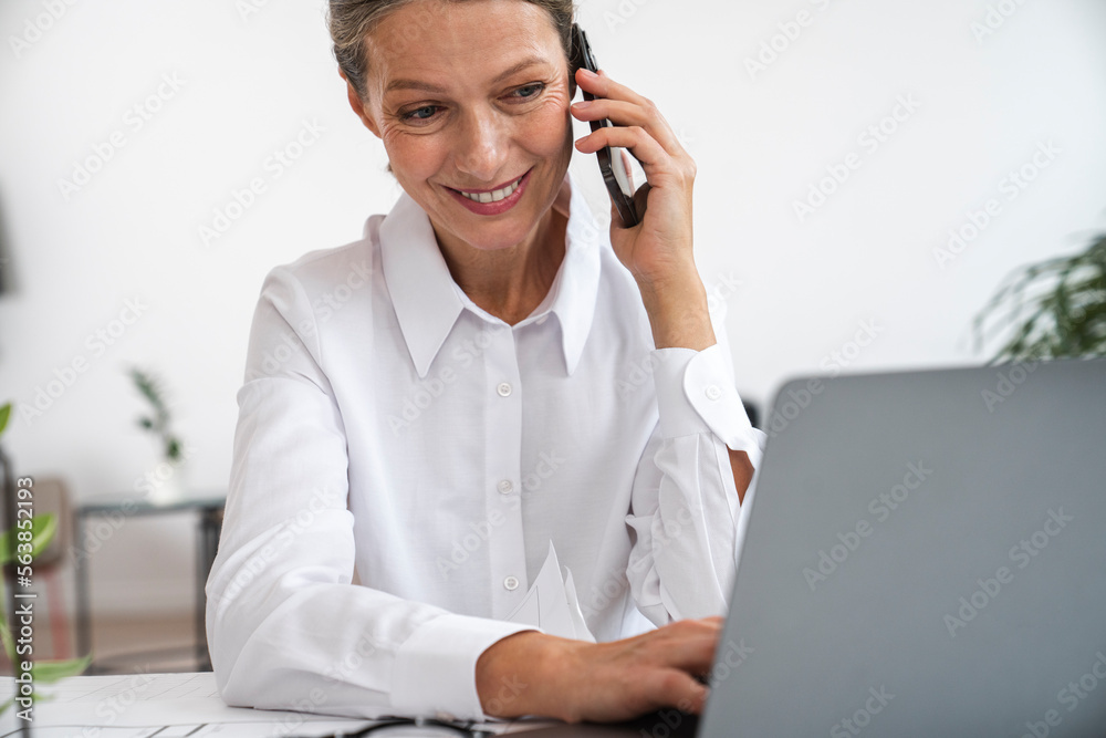 Happy mature businesswoman talking over mobile phone using laptop in office Happy mature businesswoman talking over mobile phone using laptop in office