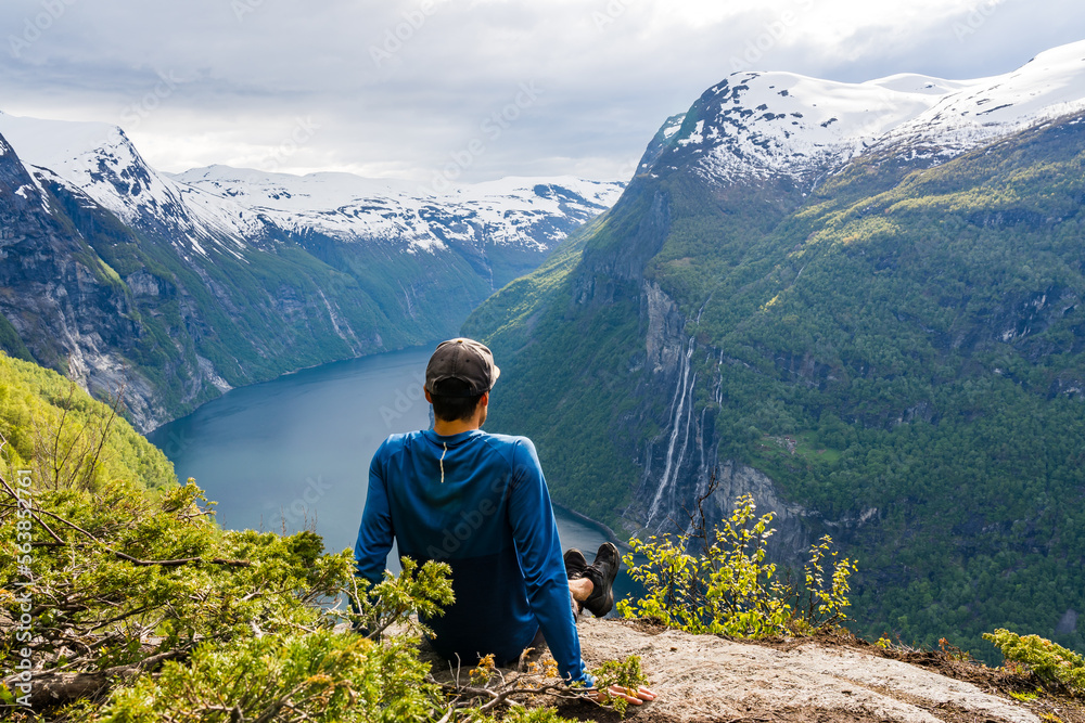Man resting on the rocks during a hike high above Geiranger fjord with snowcapped peaks and the seven sisters waterfall in the background