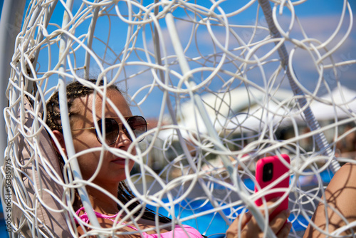 Through white weave of modern hanging chair  you can see young woman in dark sunglasses with phone. Close up of weaving of modern hanging chair on the beach and a girl sitting on soft pillows in it