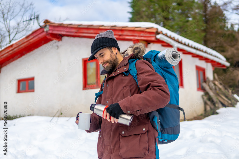 Man drinking coffee from a hot thermos in winter in the snow next to a cabin