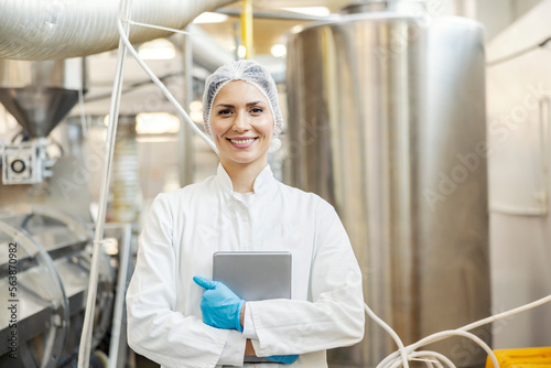 A milk processing factory worker is standing in front of the tank with milk and holding tablet in her hands.