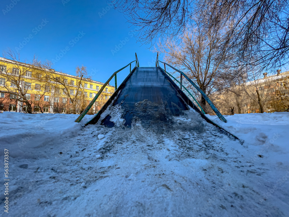 Obraz premium Children's metal winter slide standing in the yard surrounded by houses, trees and against the blue sky