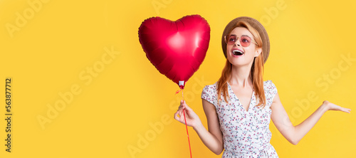 happy girl in straw hat and sunglasses hold love heart balloon on yellow background. Woman isolated face portrait, banner with copy space.