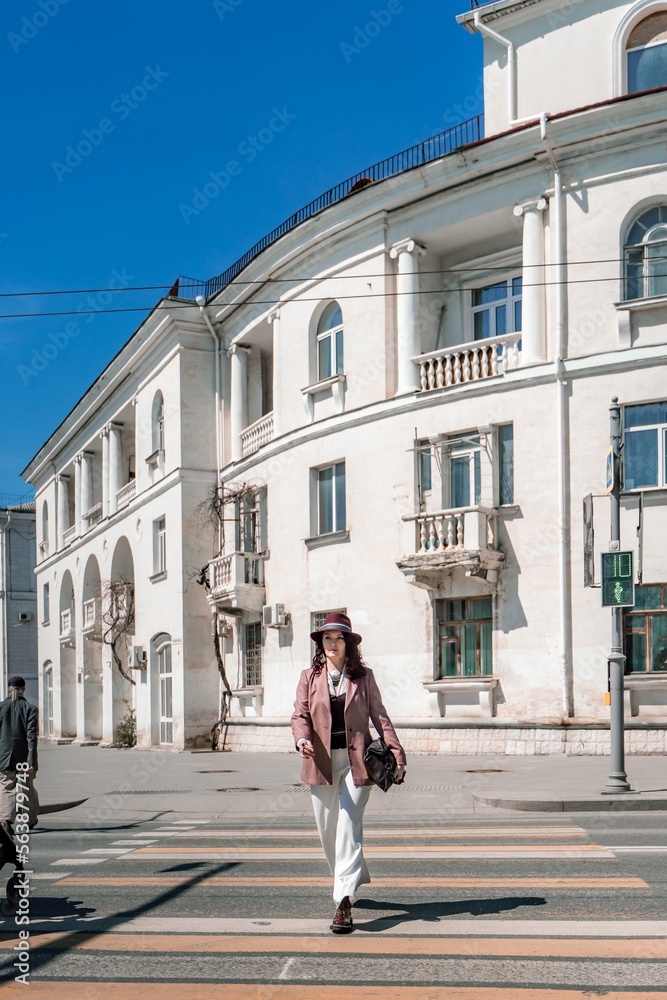 Woman city road crossing. Stylish woman in a hat crosses the road at a pedestrian crossing in the city. Dressed in white trousers and a jacket with a bag in her hands. Woman city road crossing. Stylish woman in a hat crosses the road at a pedestrian crossing in the city. Dressed in white trousers and a jacket with a bag in her hands.