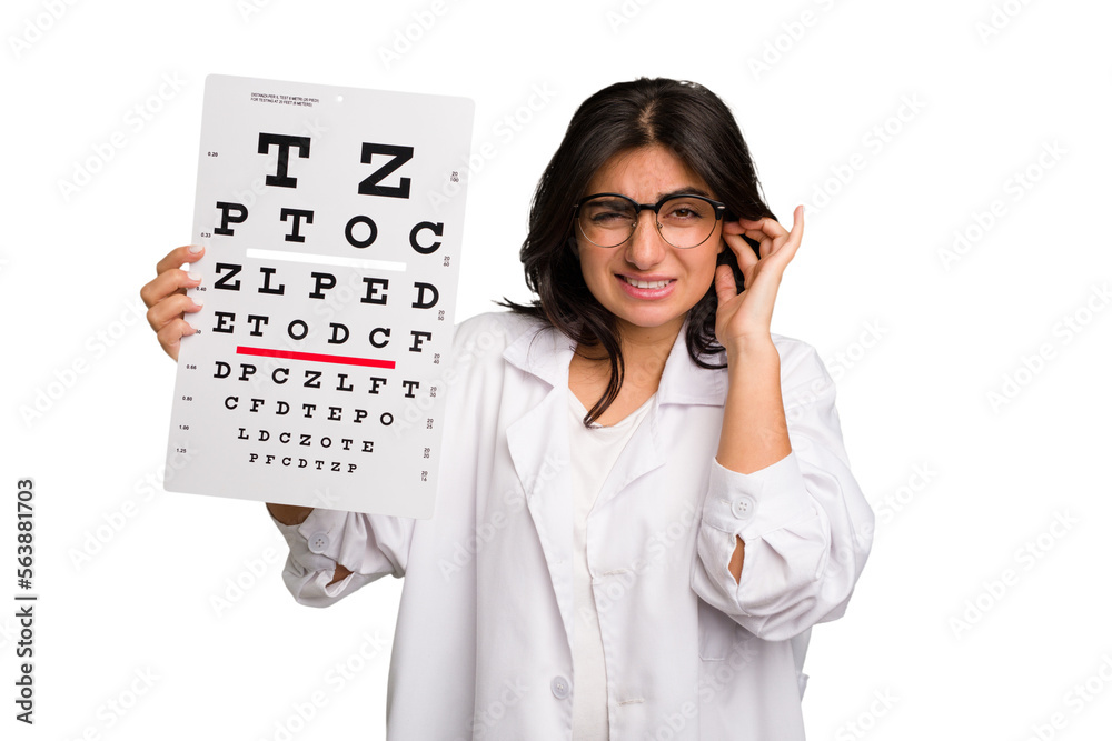 Young indian oculist woman holding an eye chart paper cut out isolated covering ears with hands. Young indian oculist woman holding an eye chart paper cut out isolated covering ears with hands.