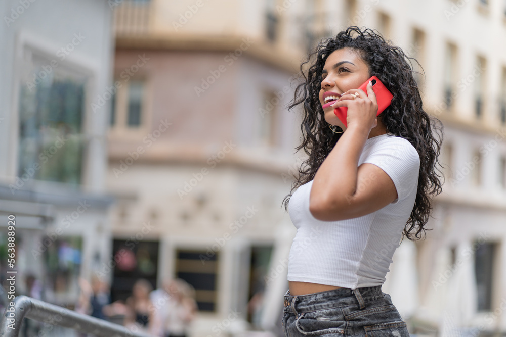 Young brunette woman talking very happy with a friend with her red mobile phone and dressed in a white t-shirt.