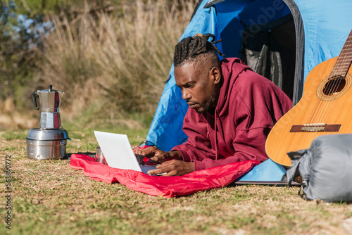 african american man lying down in camping tent using laptop computer and working remotely while camping in nature