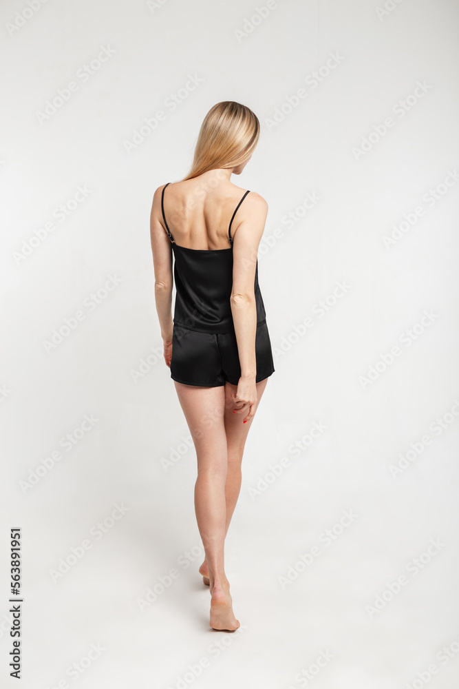 Beautiful slim young woman with sexy slender legs in a fashionable black nightgown with a tank top and shorts on a white background, back view