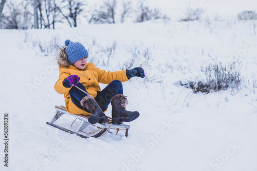 Funny child boy in colorful yellow jacket having fun sledding down the mountain on winter snowy vacation