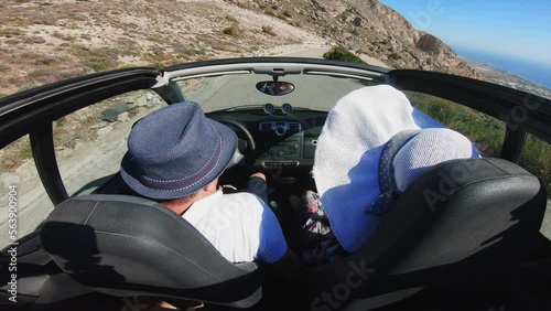 Driving along curved coastal road along Aegean sea on Santorini island at Ancient Thera, Greece. Couple with hats enjoy honneymoon photo