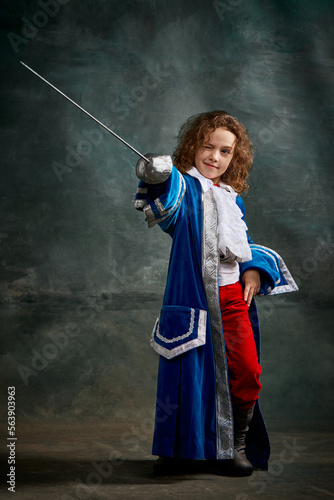 Emotional kid, little girl wearing costume of prince, musketeer and royal person posing over dark vintage style background. Fashion, theater, beauty, emotions concept