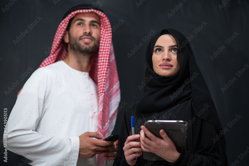 Young muslim business couple arabian man with woman in fashionable hijab dress using mobile phone and tablet computer in front of black chalkboard representing modern islam fashion technology Young muslim business couple arabian man with woman in fashionable hijab dress using mobile phone and tablet computer in front of black chalkboard representing modern islam fashion technology