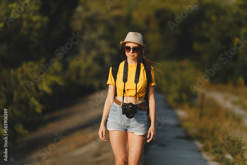 Young attractive travel photographer girl in hat, yellow t-shirt, denim shorts, sunglasses, with photo camera smiling and hiking  © Natalia