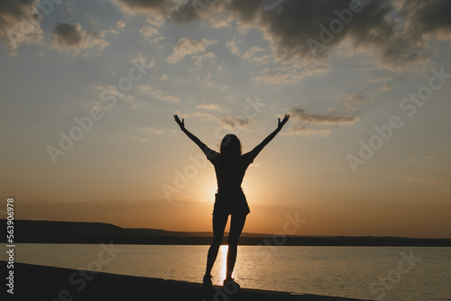 The silhouette of young sexy girl enjoying the sundown with raised up hands at the Italian lake beach 