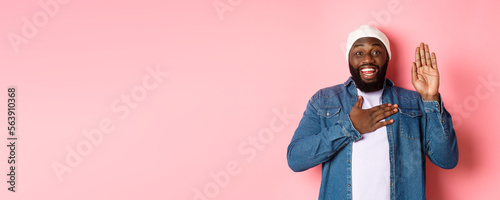 Image of smiling african-american man being honest, telling truth, hands on heart and in air, making promise or swearing, standing ove rpink background photo