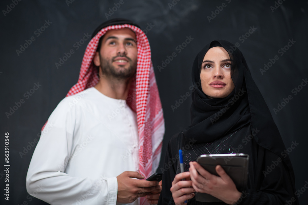 Young muslim business couple arabian man with woman in fashionable hijab dress using mobile phone and tablet computer in front of black chalkboard representing modern islam fashion technology 