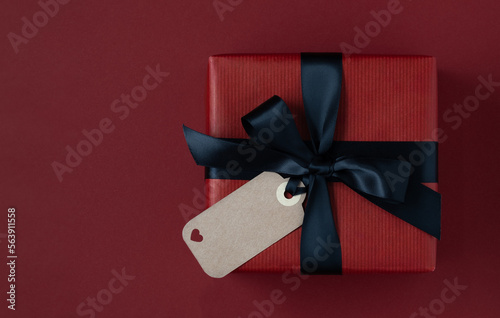 Red gift box with black ribbon and empty craft gift tag on red background. Space for text.