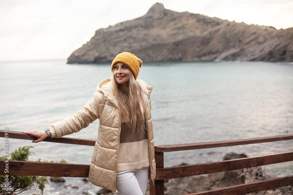 Fototapeta premium A beautiful woman walks on the beach in cold weather, meditates to the sound of the waves. The girl dressed sweater, white jeans and yellow hat