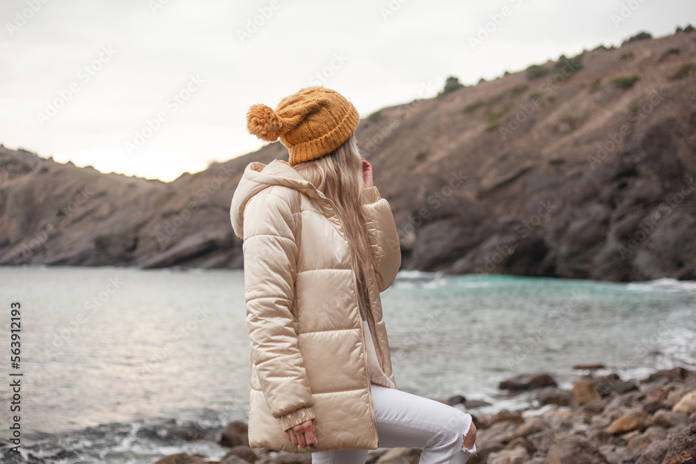 A beautiful woman walks on the beach in cold weather, meditates to the sound of the waves. The girl dressed sweater, white jeans and yellow hat