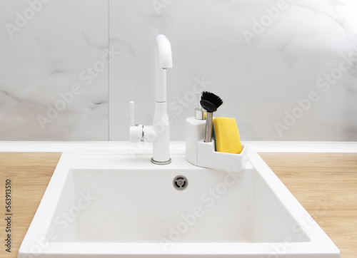 Beautiful kitchen interior of a white sink with a water faucet, sponge and brush for washing dishes and detergent against a white tile wall. Modern interior
